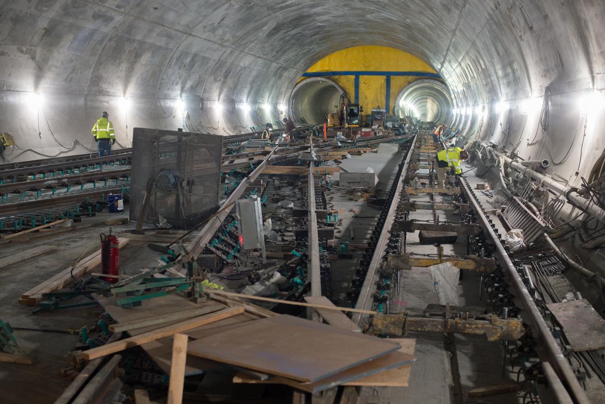 Workers in safety vests construct track infrastructure inside a subway tunnel with curved walls and overhead lighting.