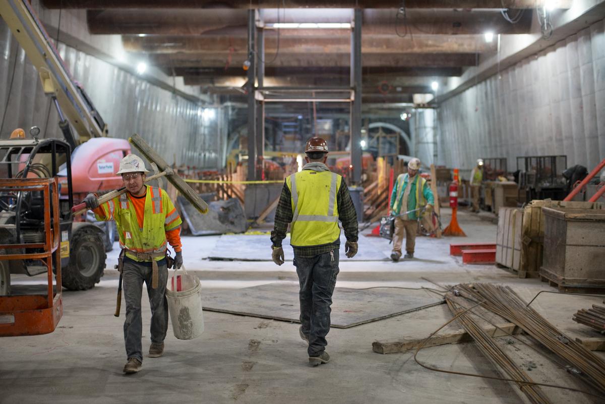 Construction workers in safety vests and hard hats walk through an industrial tunnel with equipment and building materials.