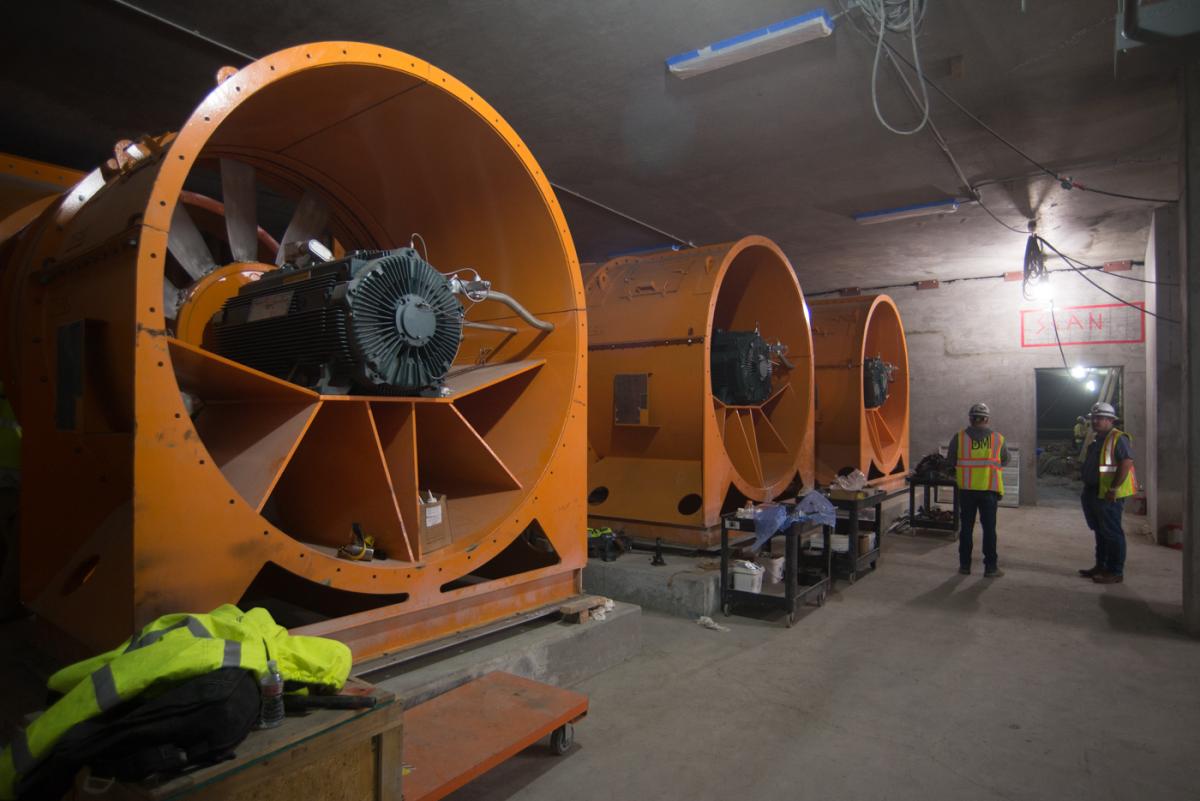 Large orange ventilation fans installed in underground station construction area with workers in safety vests.