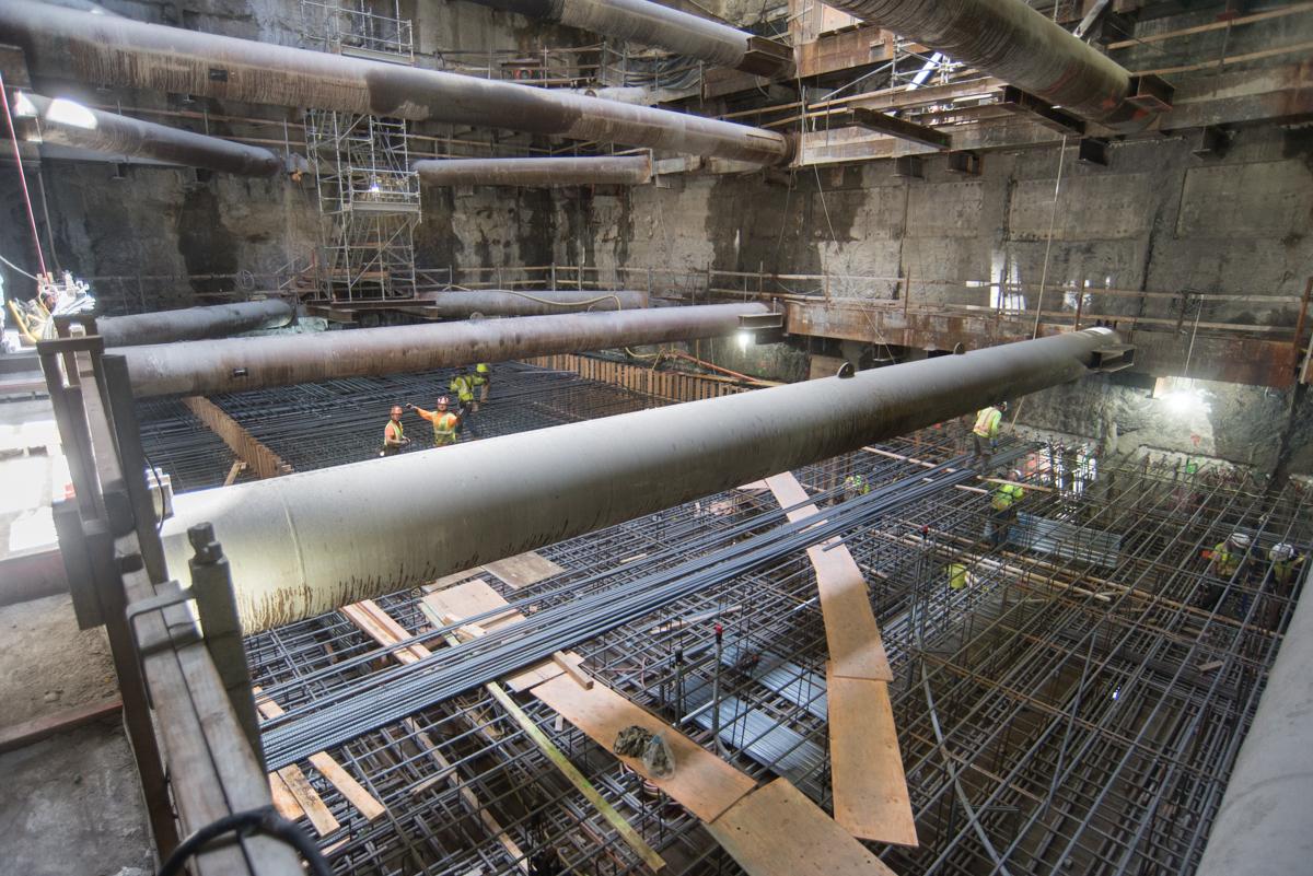 Construction workers assemble rebar and large pipes in underground transit tunnel cavern with concrete walls and scaffolding.