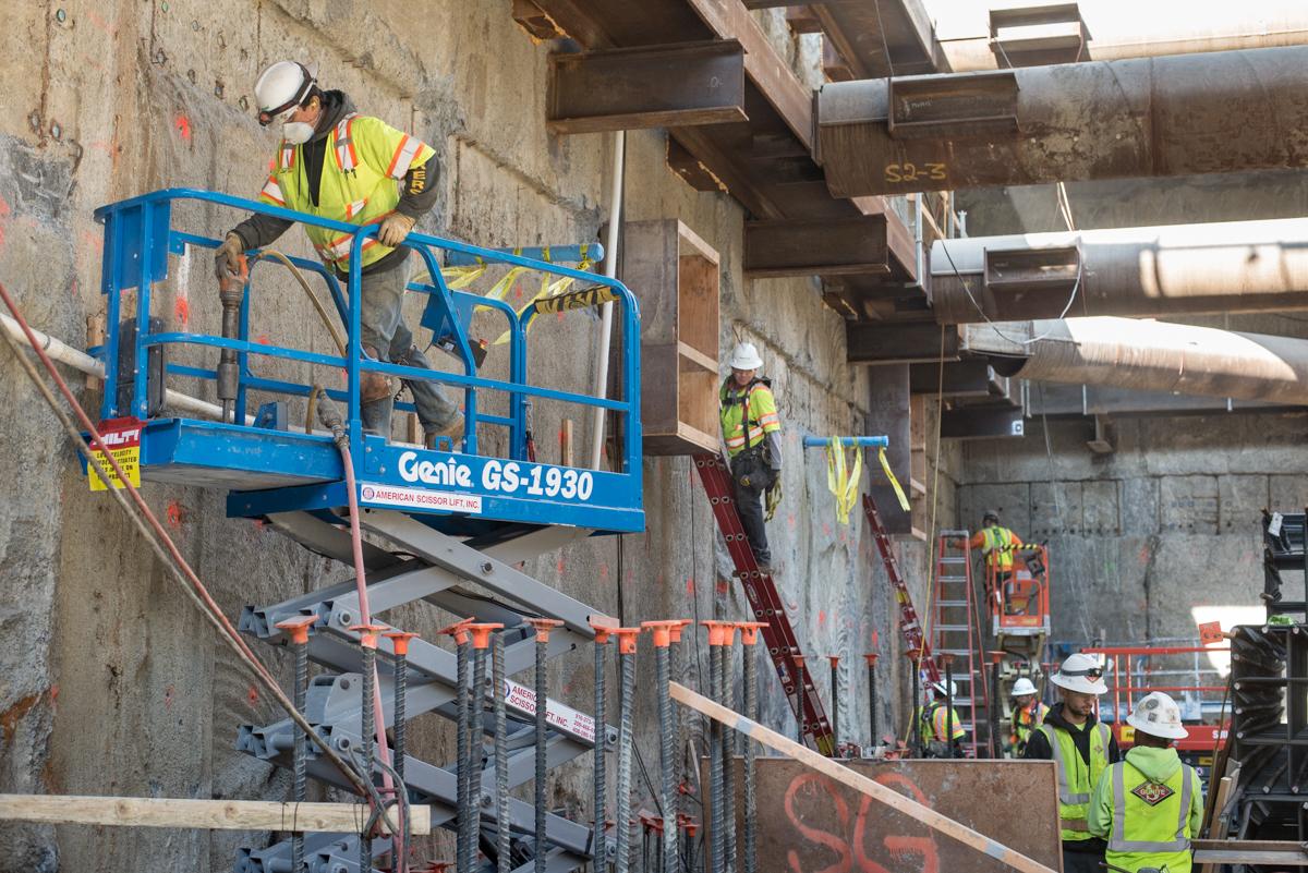 Construction workers on a blue Genie lift working on concrete walls at a Central Subway station construction site.