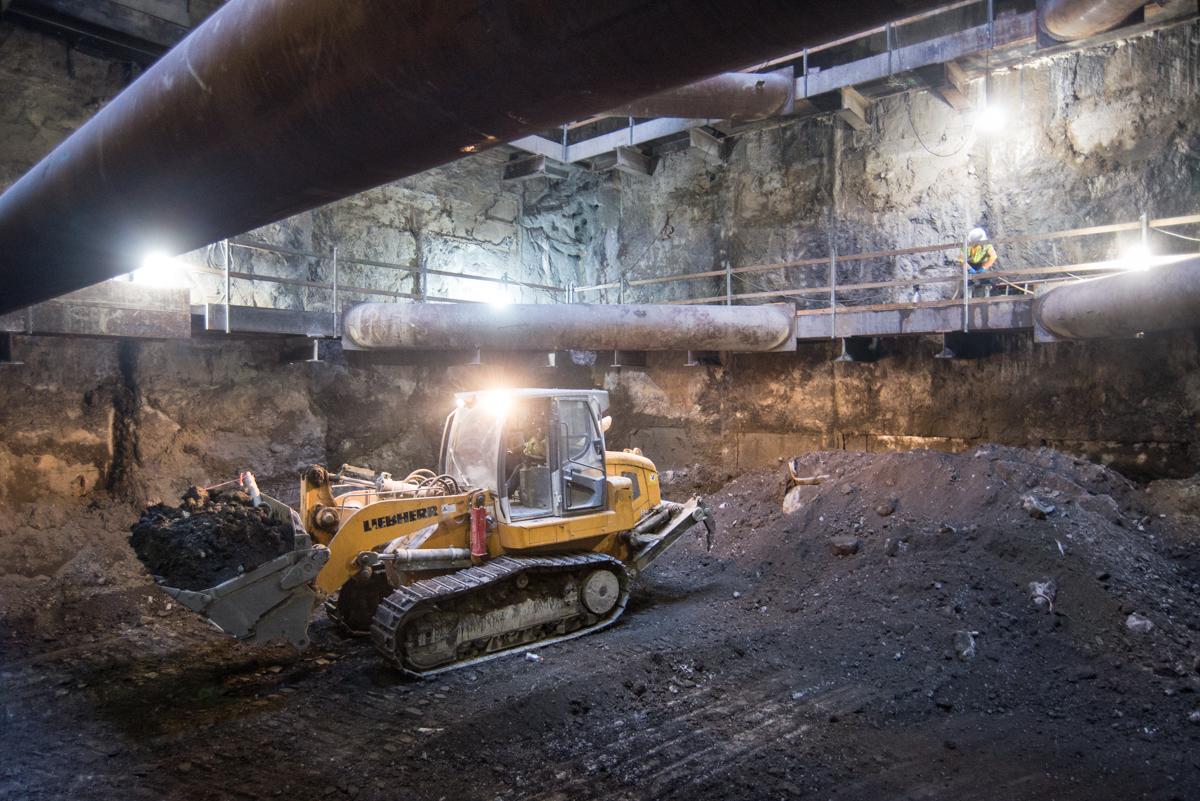 Yellow bulldozer excavating material in underground station cavern with concrete walls and overhead pipes.