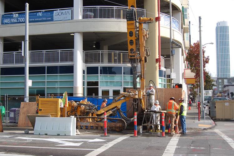 Construction crew operating drilling equipment on 4th Street with traffic controls and barriers in place.