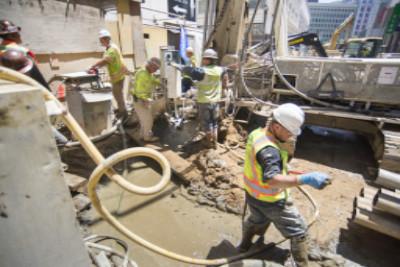 Construction workers in safety gear operating concrete pump equipment on an urban street work site.