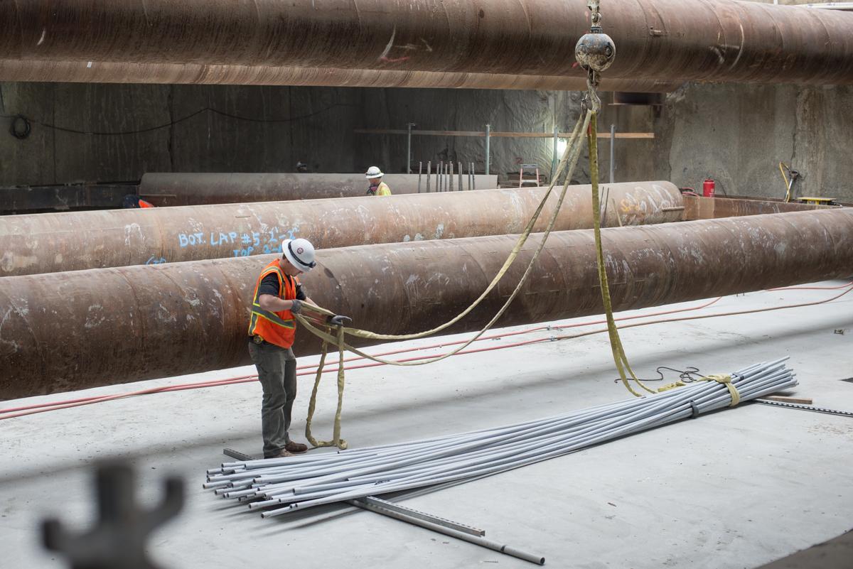 Worker in safety gear prepares rigging to lift bundle of PVC conduit with crane at transit station construction site.