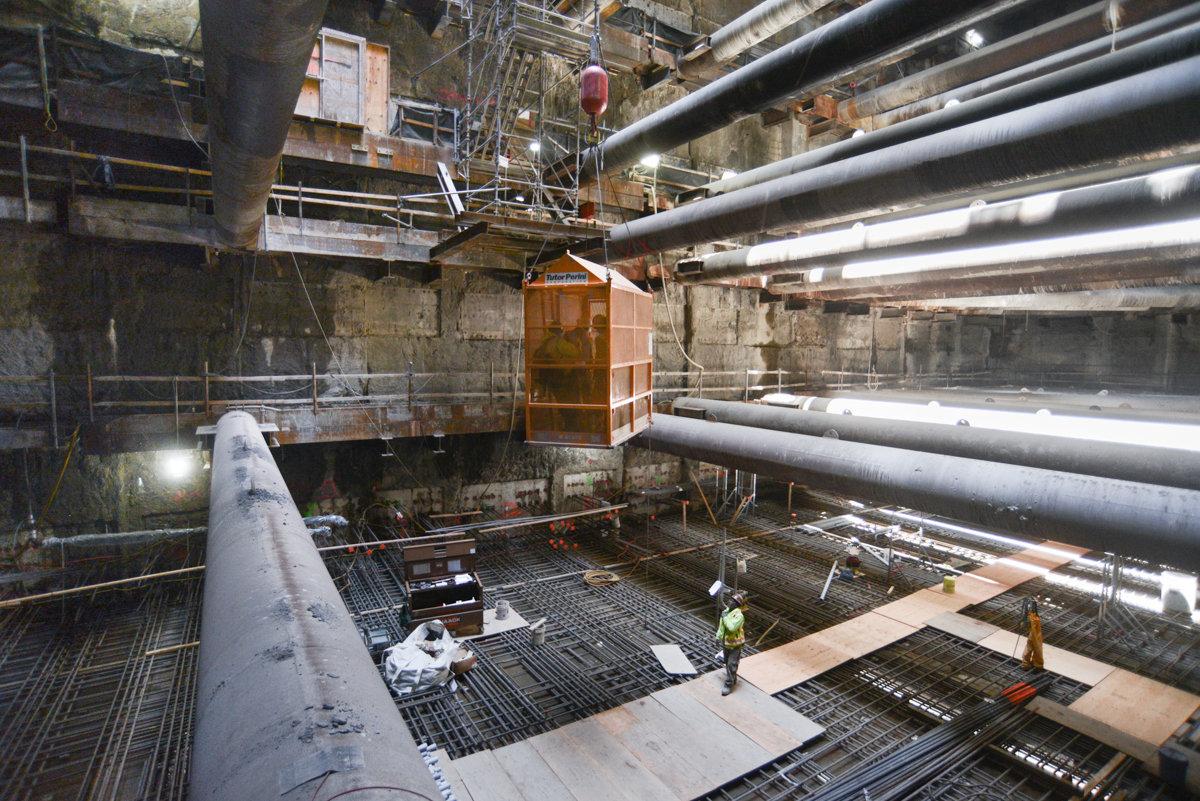 Workers in underground station construction pit with large pipes and equipment overhead.