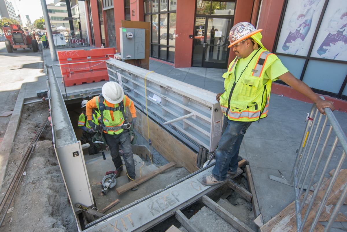 Two construction workers in safety gear work on an underground staircase structure at a street-level construction site.