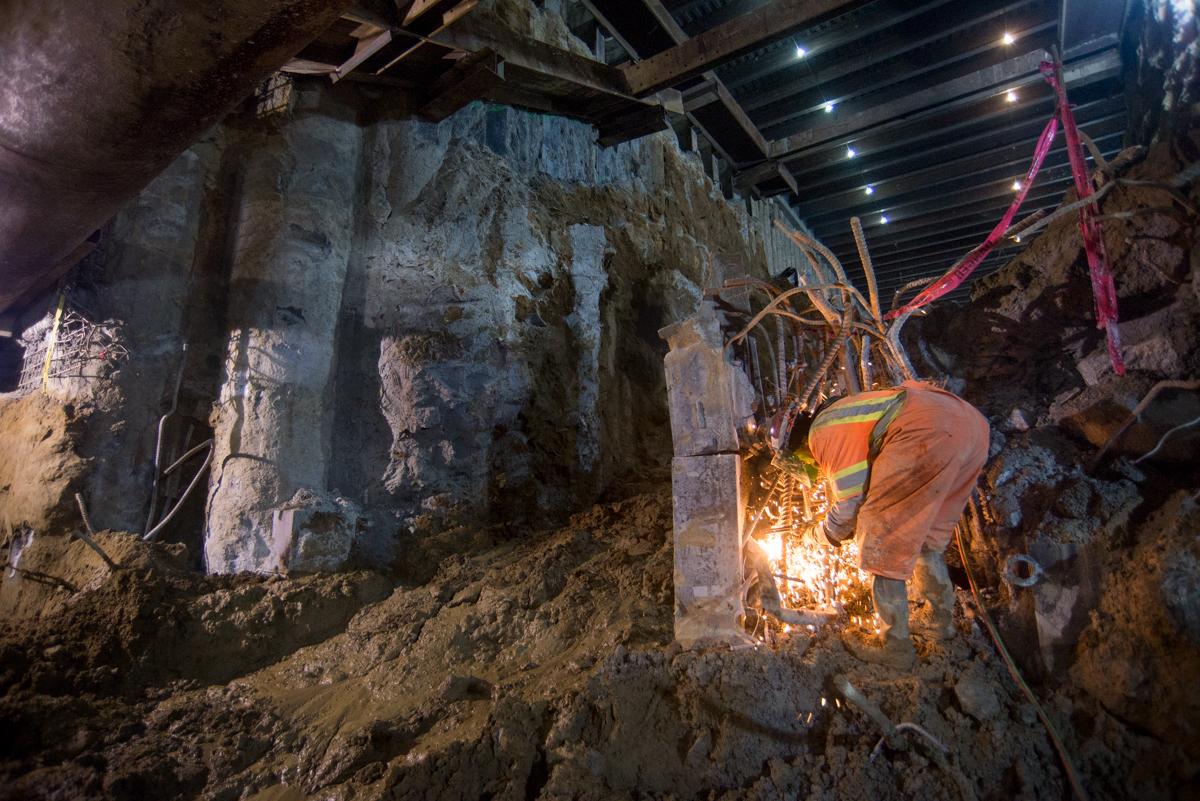 Worker in orange safety vest welding steel reinforcement at underground transit construction site.
