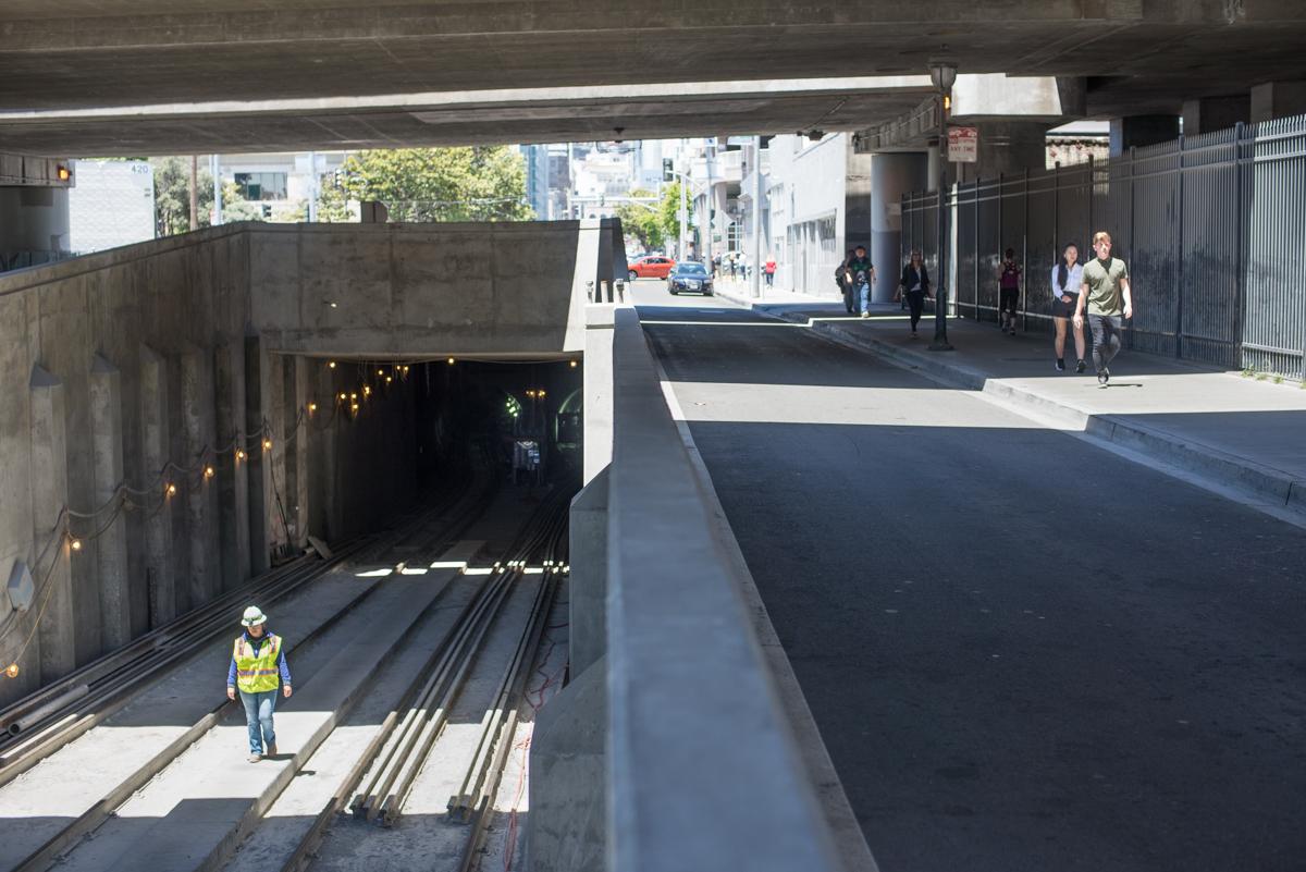 Worker in safety vest stands in tunnel entrance with railroad tracks visible; pedestrians walk on street level above.
