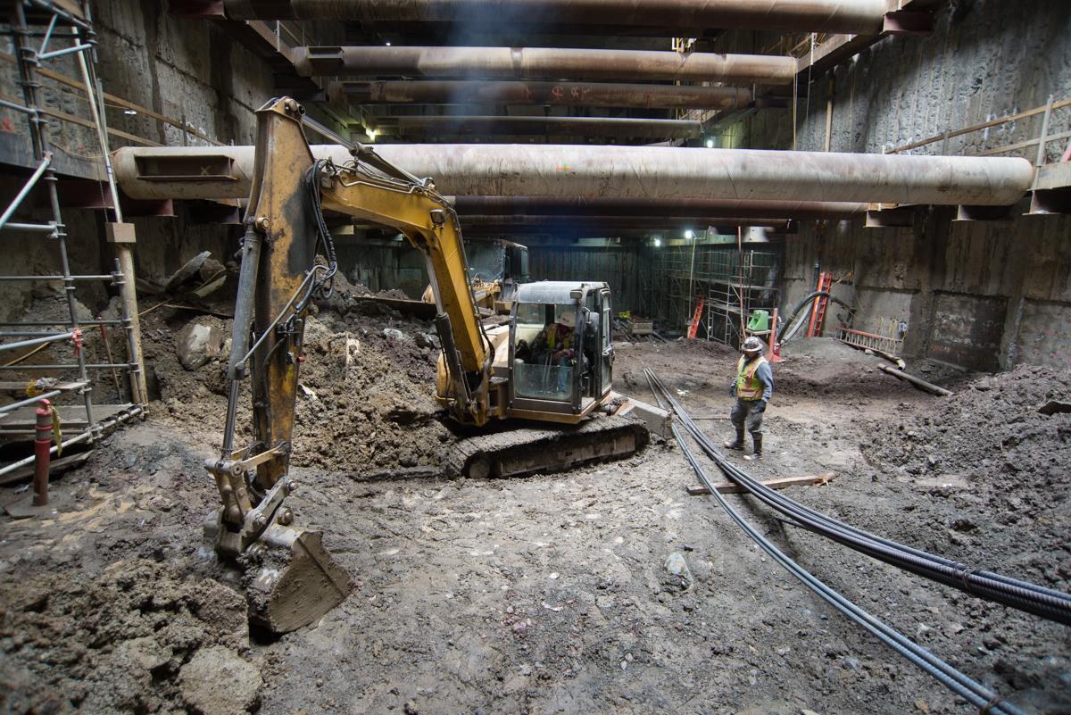 Excavator removing material from underground station headhouse construction site with workers and temporary steel supports.