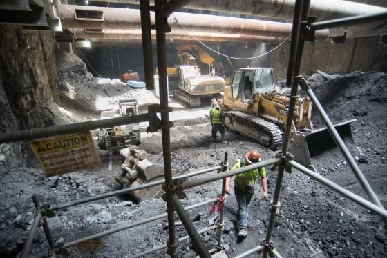 Construction workers in safety vests operate excavation equipment in underground Chinatown Station headhouse.