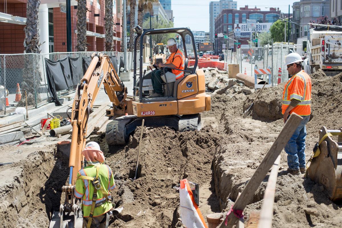 Construction crew excavating utility trench on 4th Street with backhoe and hand tools.