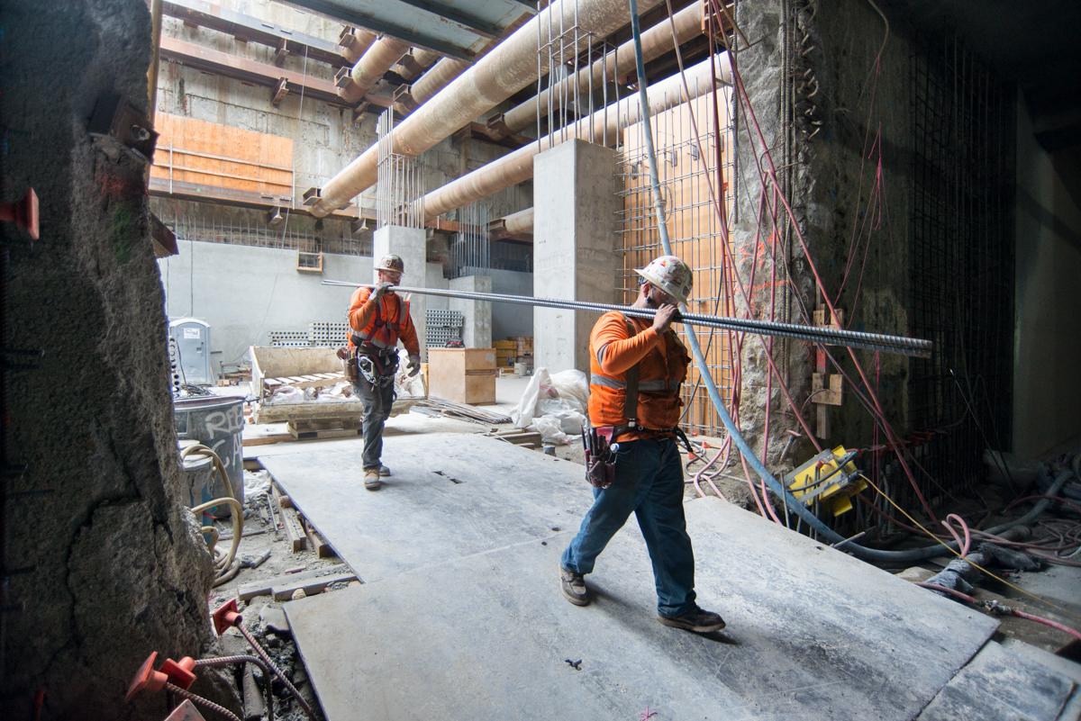 Two construction workers in safety gear carrying rebar inside an underground transit station under construction.