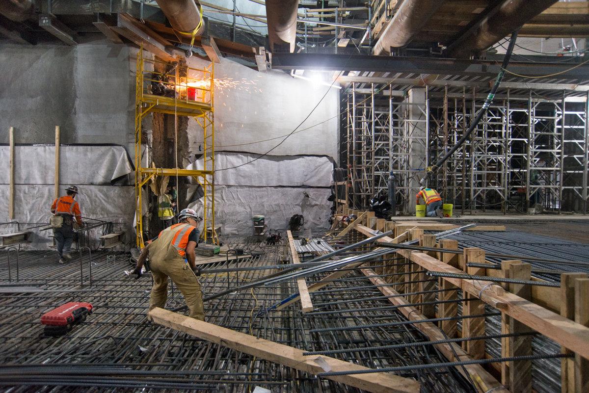 Construction workers install rebar and utilities in underground transit station concourse during active building work.