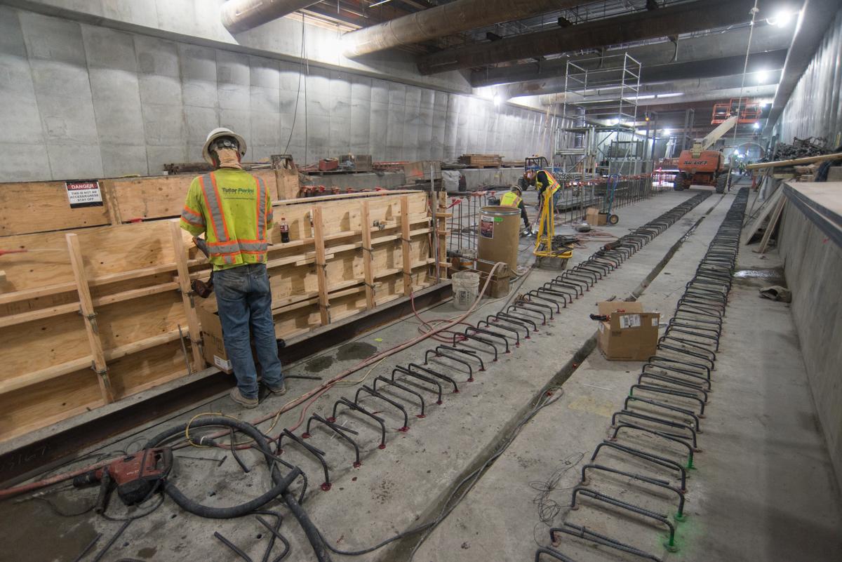 Workers in safety vests install reinforcing steel in concrete forms for transit station platform construction.