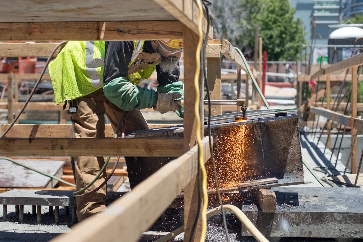 Construction worker using cutting torch to fabricate metal brackets on building structure at transit station.