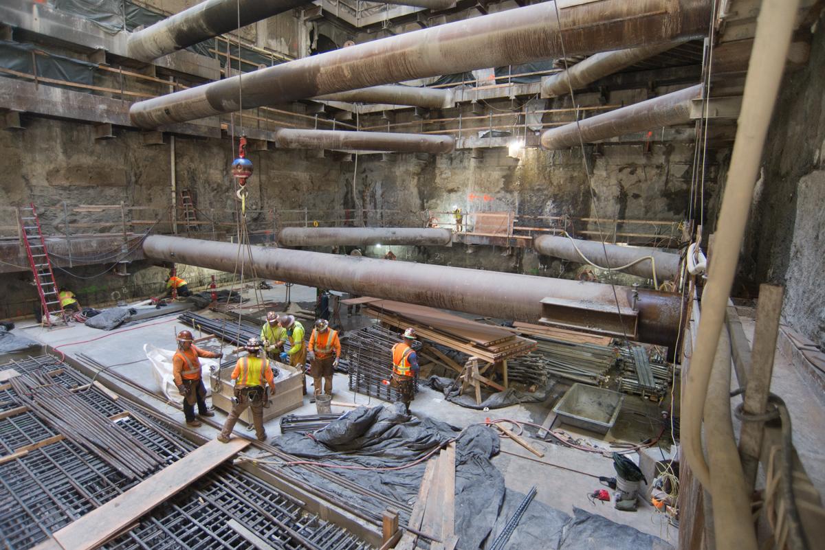 Construction workers in safety gear inside underground transit station with exposed pipes and reinforced concrete structure.