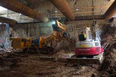 Heavy equipment including a yellow drill rig and red and white excavator inside an underground construction site with concrete pillars and dirt floor.