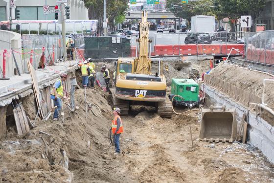 Excavation work on 4th Street with CAT equipment, workers in safety gear, and construction barriers.
