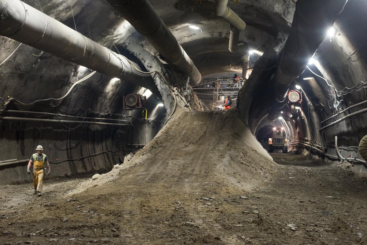 Worker in hard hat walks past large pile of excavated soil in underground tunnel with overhead pipes and lighting.