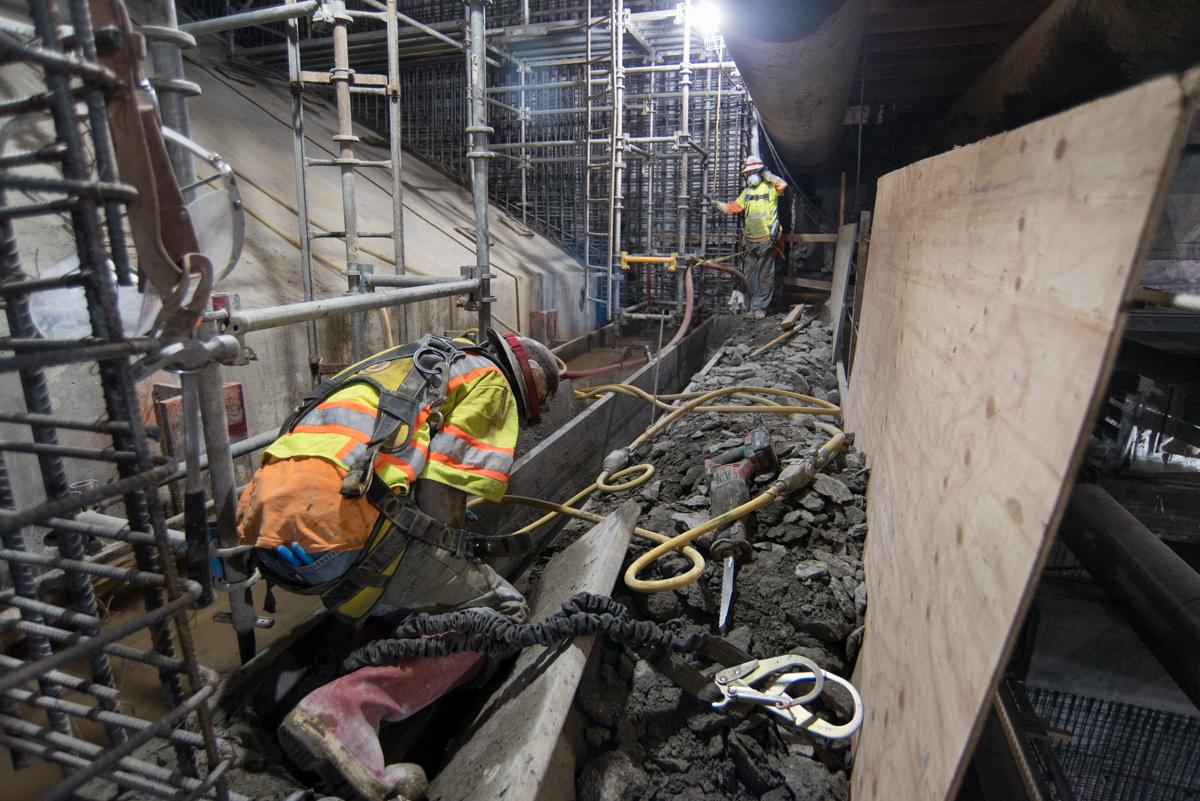 Workers in safety vests remove concrete debris from underground construction site with temporary bracing and utilities visible.