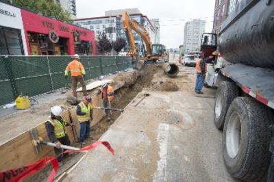 Construction workers install sewer line on urban street with heavy equipment and trucks.