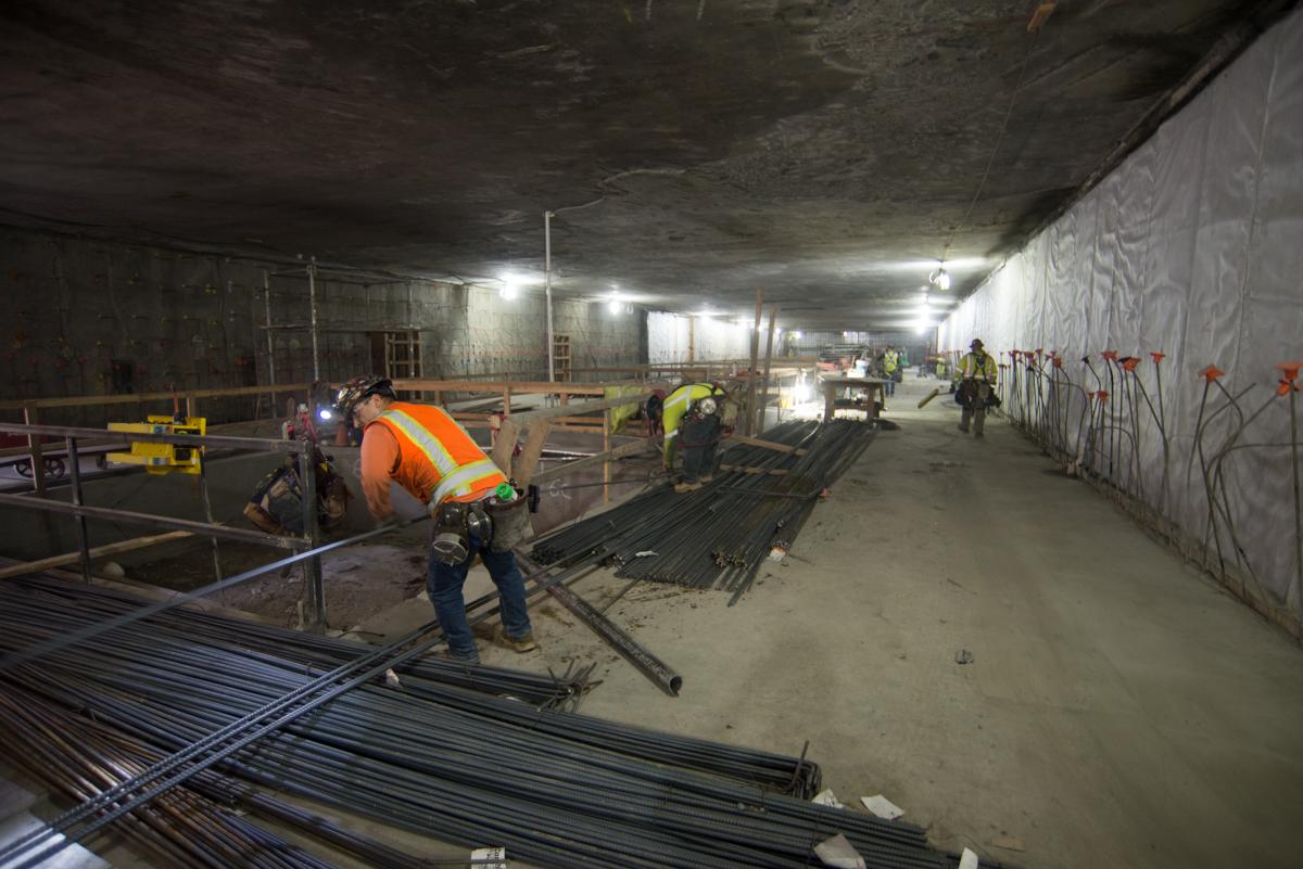 Workers in safety vests install rebar and support structures in underground transit station construction site.