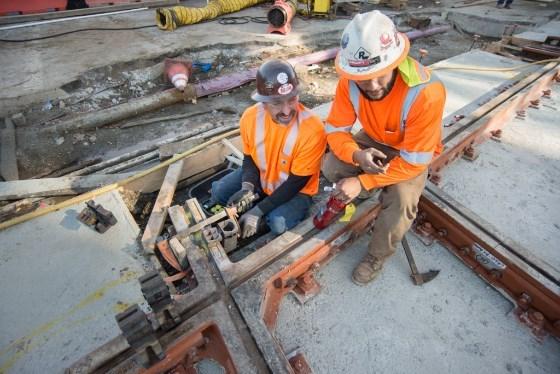 Two workers in orange safety vests and hard hats prepare welding equipment on subway track infrastructure.