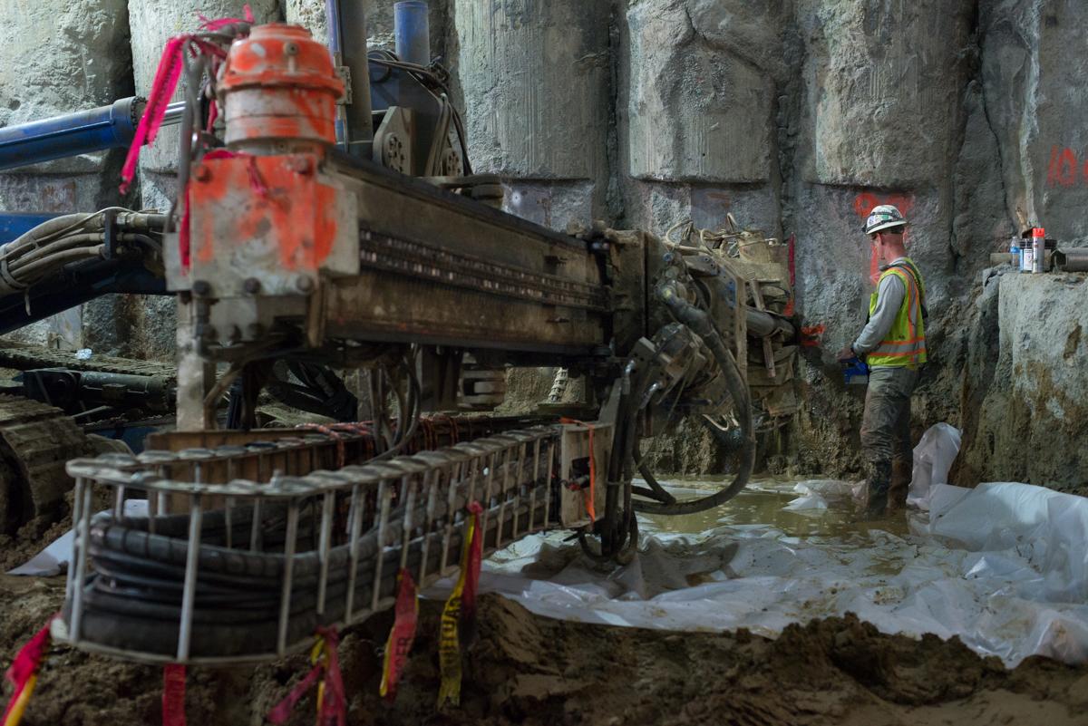 Construction worker in safety gear stands near excavation equipment and temporary bracing at underground transit station site.