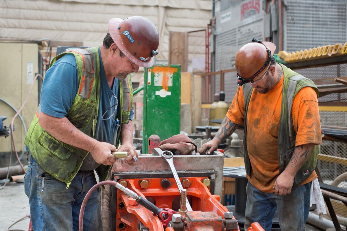 Two workers in safety gear operate a jackhammer on an orange machine in an industrial workshop setting.