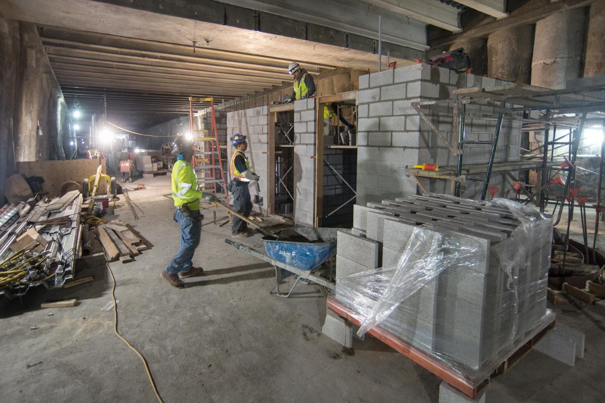 Construction workers in safety vests assemble cinderblock walls on a mezzanine level, with materials and equipment visible throughout the space.