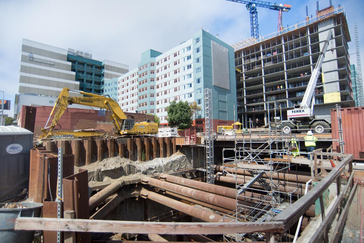 Construction excavation site with yellow machinery, steel pilings, and multi-story buildings under development in urban area.