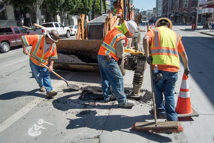 Workers in safety vests patch asphalt on a city street using equipment and tools.