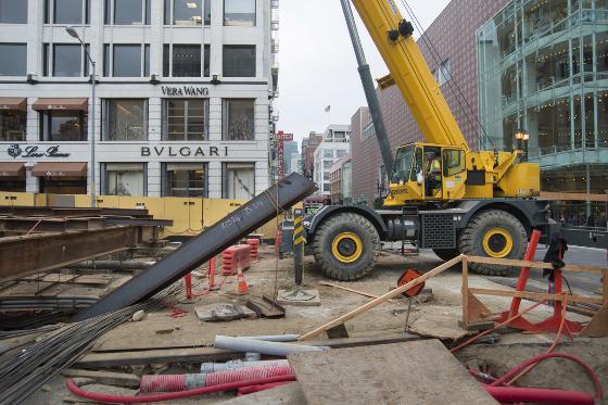 Yellow telescopic crane lifting large pipe during street construction work in urban area