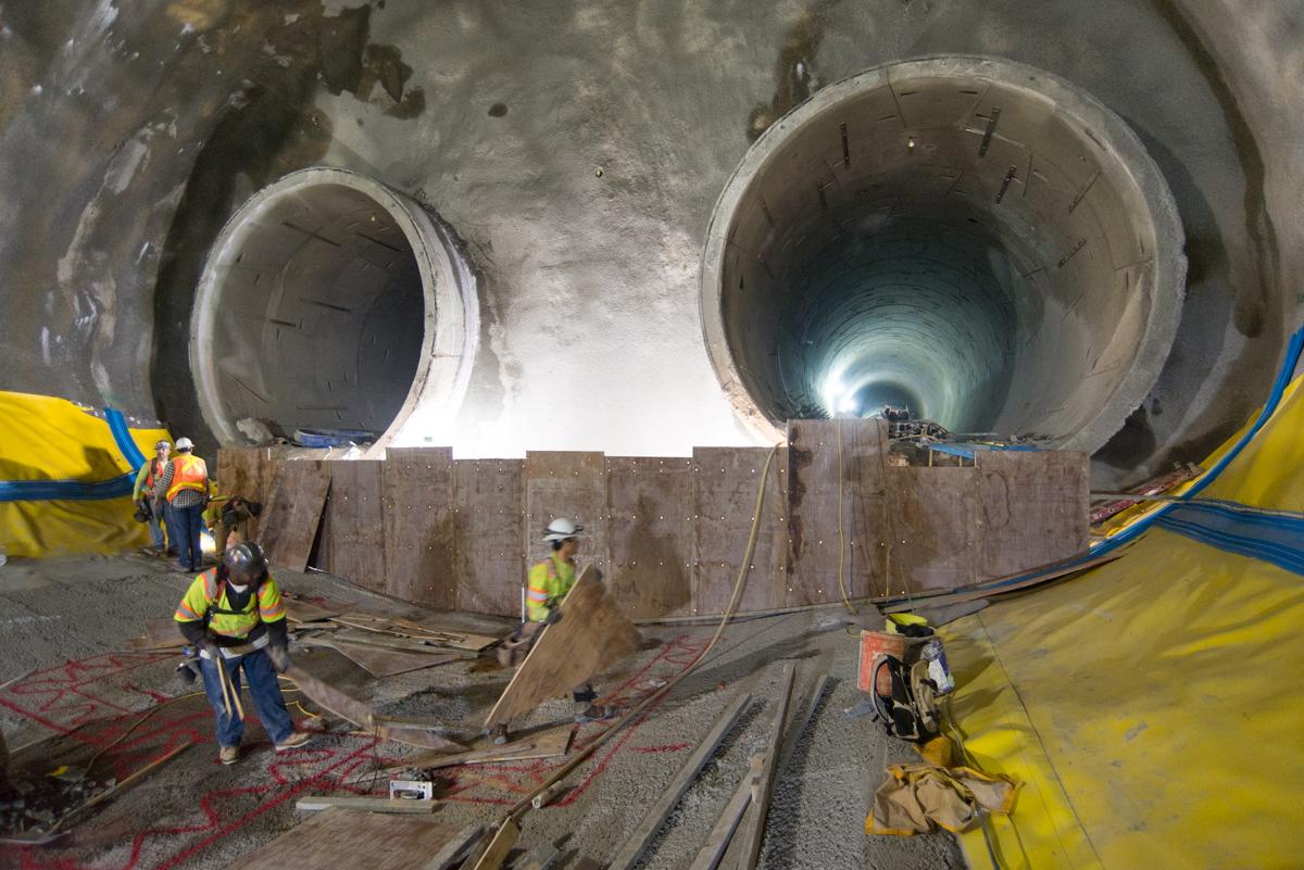 Workers in safety gear stand before two large tunnel sections during underground transit construction at Chinatown Station.