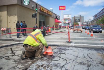 Worker in yellow safety vest crouches near street construction site with traffic cones and barriers visible.
