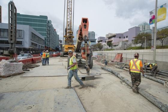 Construction workers in safety vests operate heavy machinery on 4th Street to build underground station walls.
