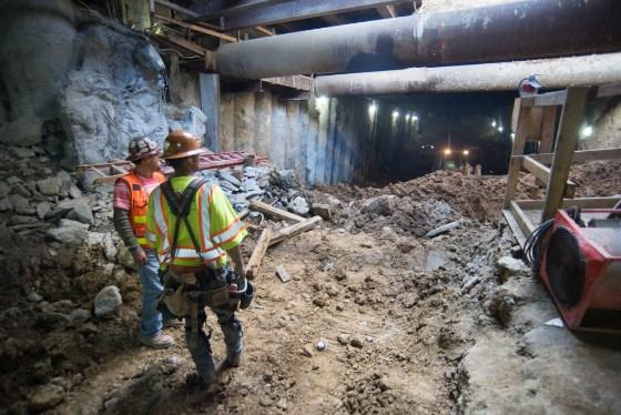 Two workers in safety vests inspect underground excavation site beneath Stockton Street with large pipes overhead.