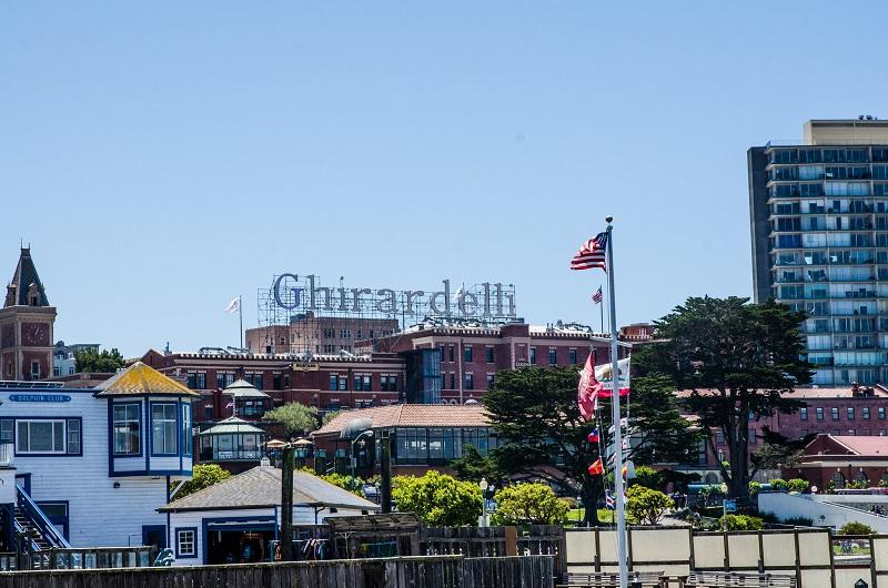 Ghirardelli Square waterfront with red brick buildings, American flag, and modern high-rises under clear blue sky.