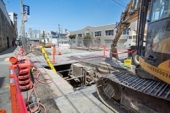Construction site with excavation pit, heavy equipment, and safety barriers on urban street.