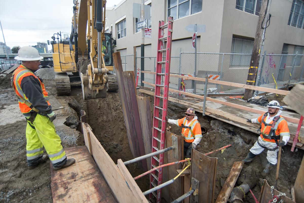 Workers in safety gear install steel sheet piles in an excavated trench at a San Francisco construction site.
