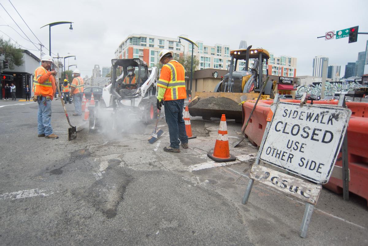 Construction crew operates equipment to remove asphalt on city street with sidewalk closure sign visible.
