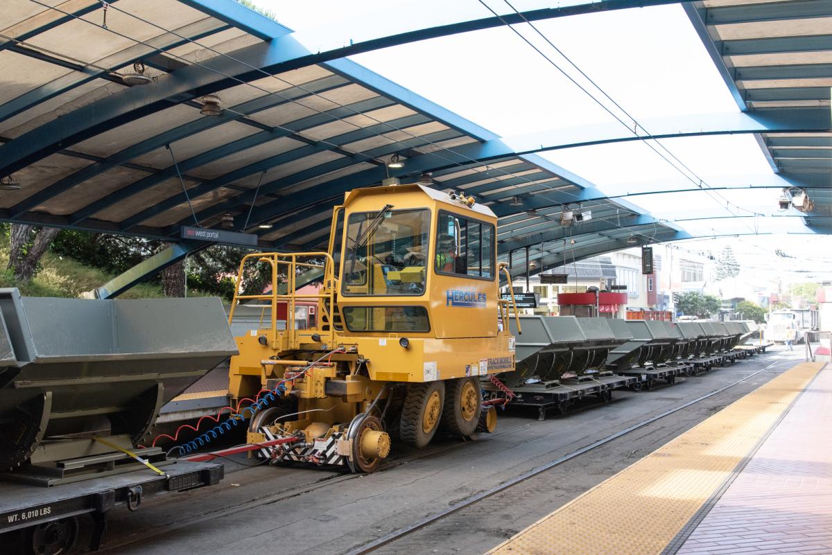 Yellow work train vehicle on tracks under covered station platform with attached cargo cars.