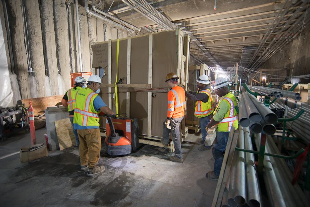 Construction crew in safety vests moving large crate with interior materials in transit station warehouse.