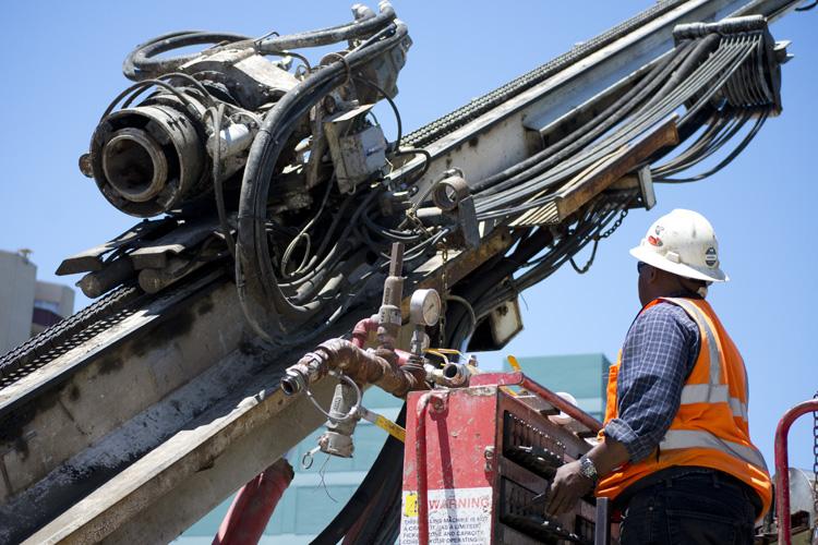 Worker in safety gear operating ground stabilization drilling equipment on urban street.