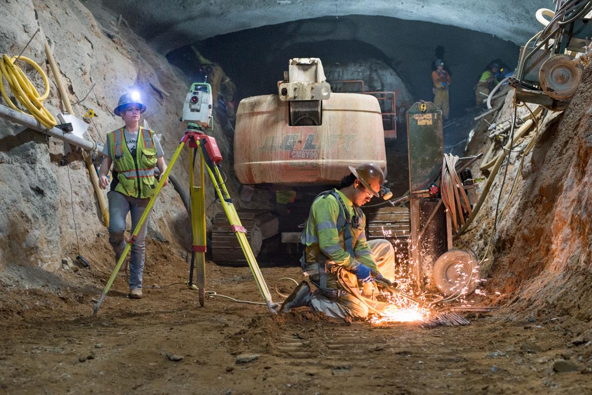 Workers in safety vests perform welding and construction inside an underground tunnel during transit infrastructure work.