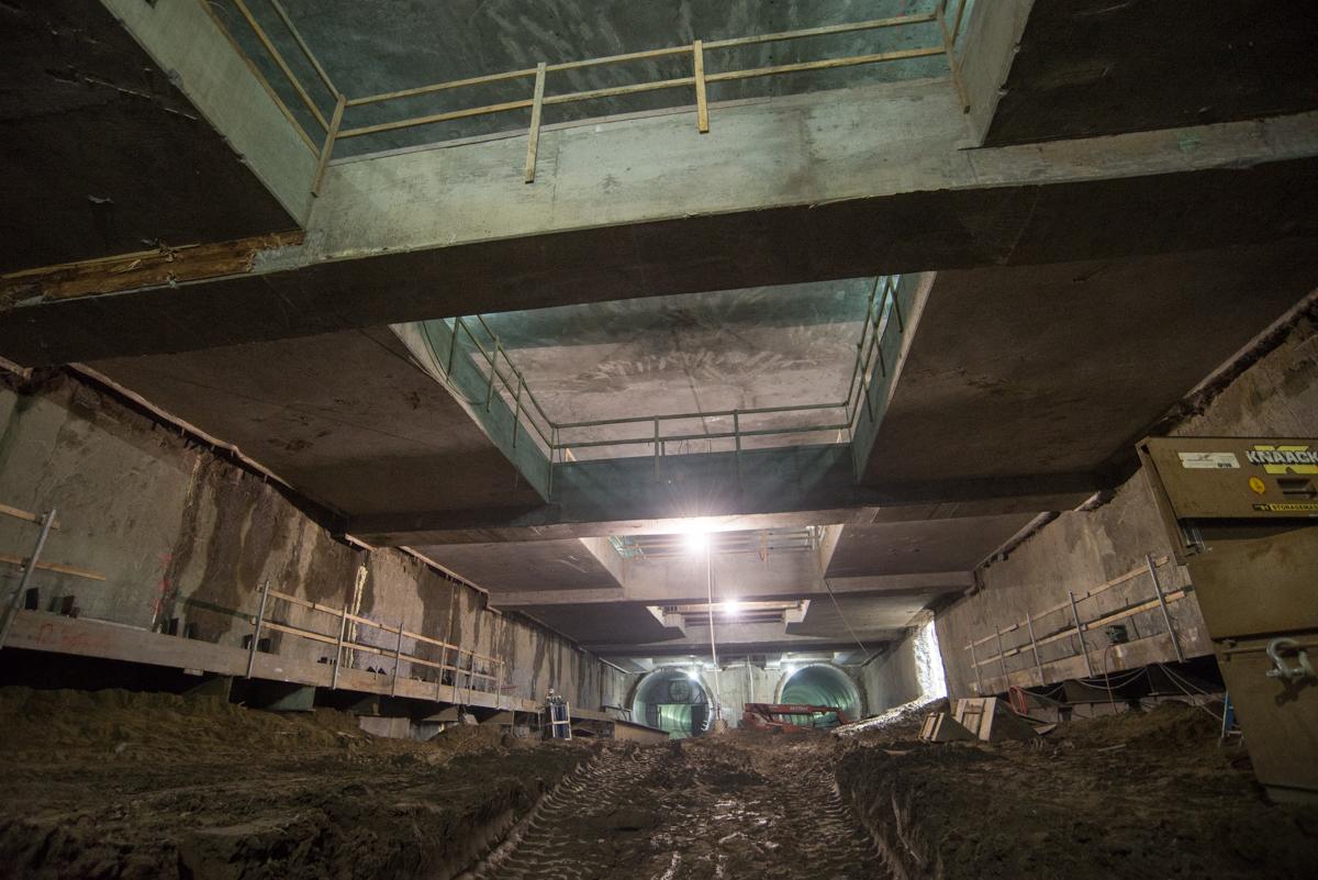 Underground transit station under construction with concrete structural elements, temporary bracing, and work lights illuminating the excavation area.