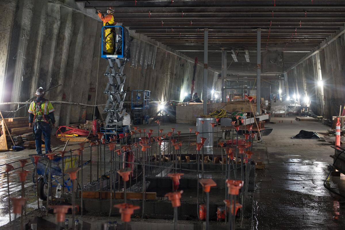 Workers on elevated lift platform inside concrete station construction site with rebar and safety barriers.