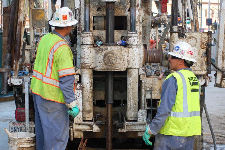 Two workers in safety vests and hard hats operate drilling equipment for subway station construction.