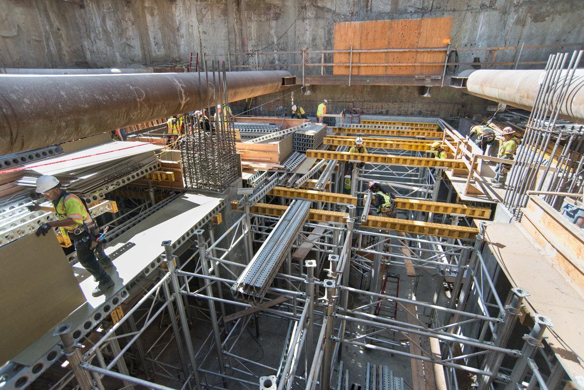 Construction workers assemble steel scaffolding and yellow support beams inside an underground transit station.